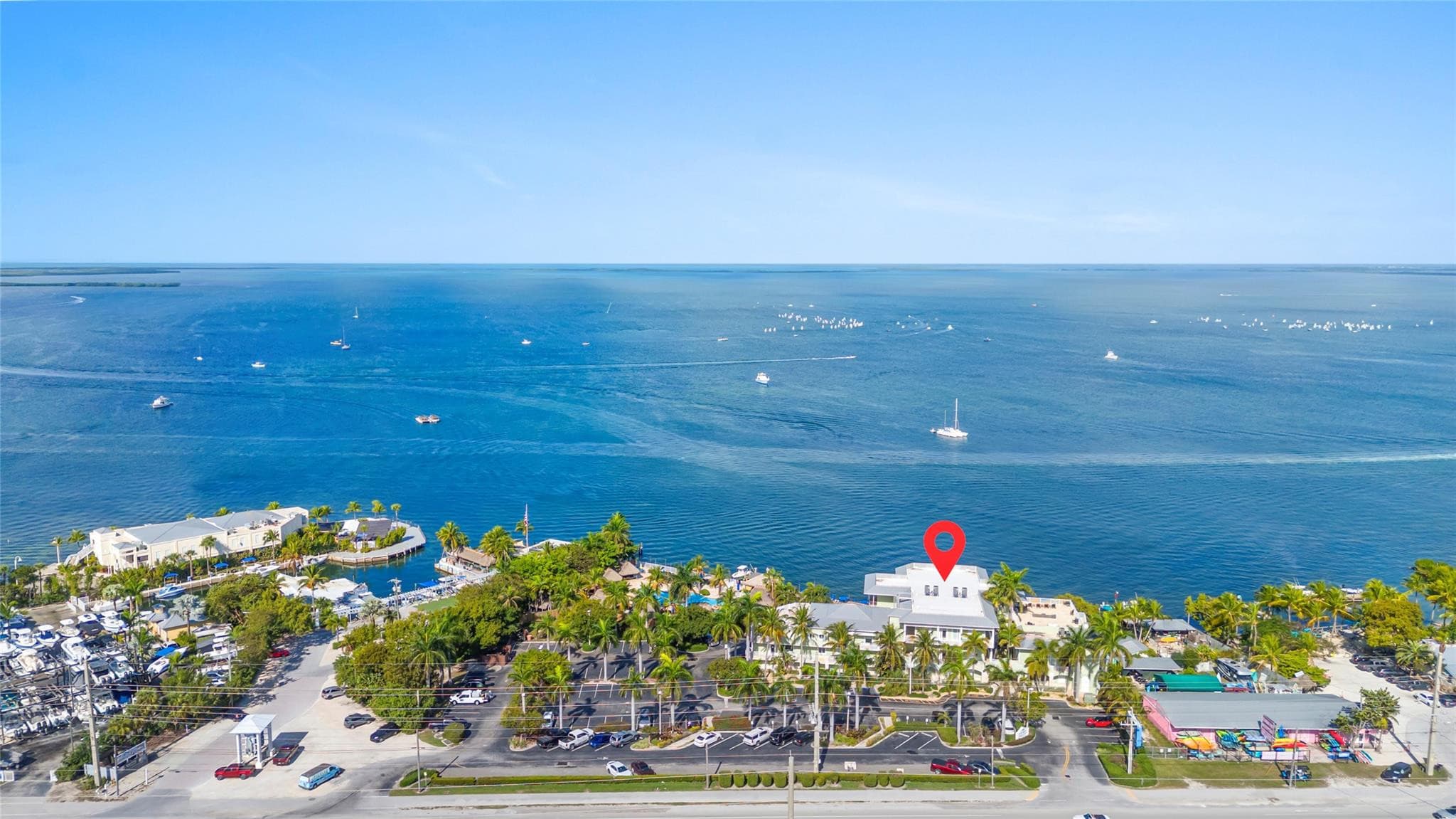 High aerial over the vast blue bay with sailboats, a red location pin marking the white-roofed bayfront residence building amid palms, docks, and the pool below.