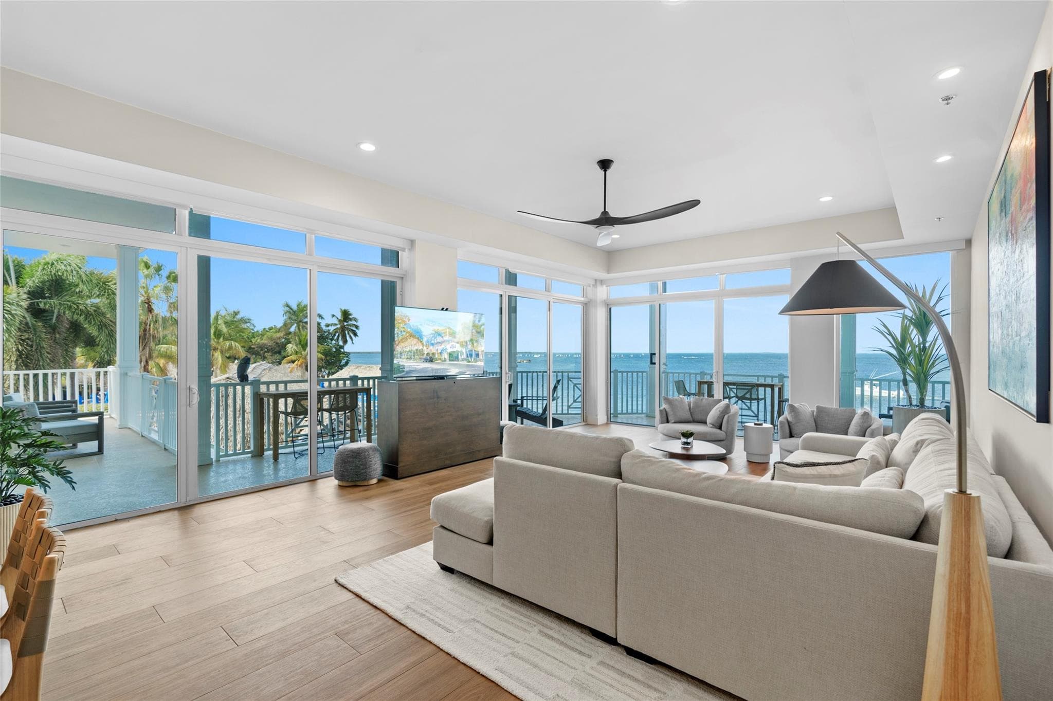 Corner living room wrapped in floor-to-ceiling windows and balcony doors with panoramic bay views, a beige sectional, arc floor lamp, and pop-up TV console.