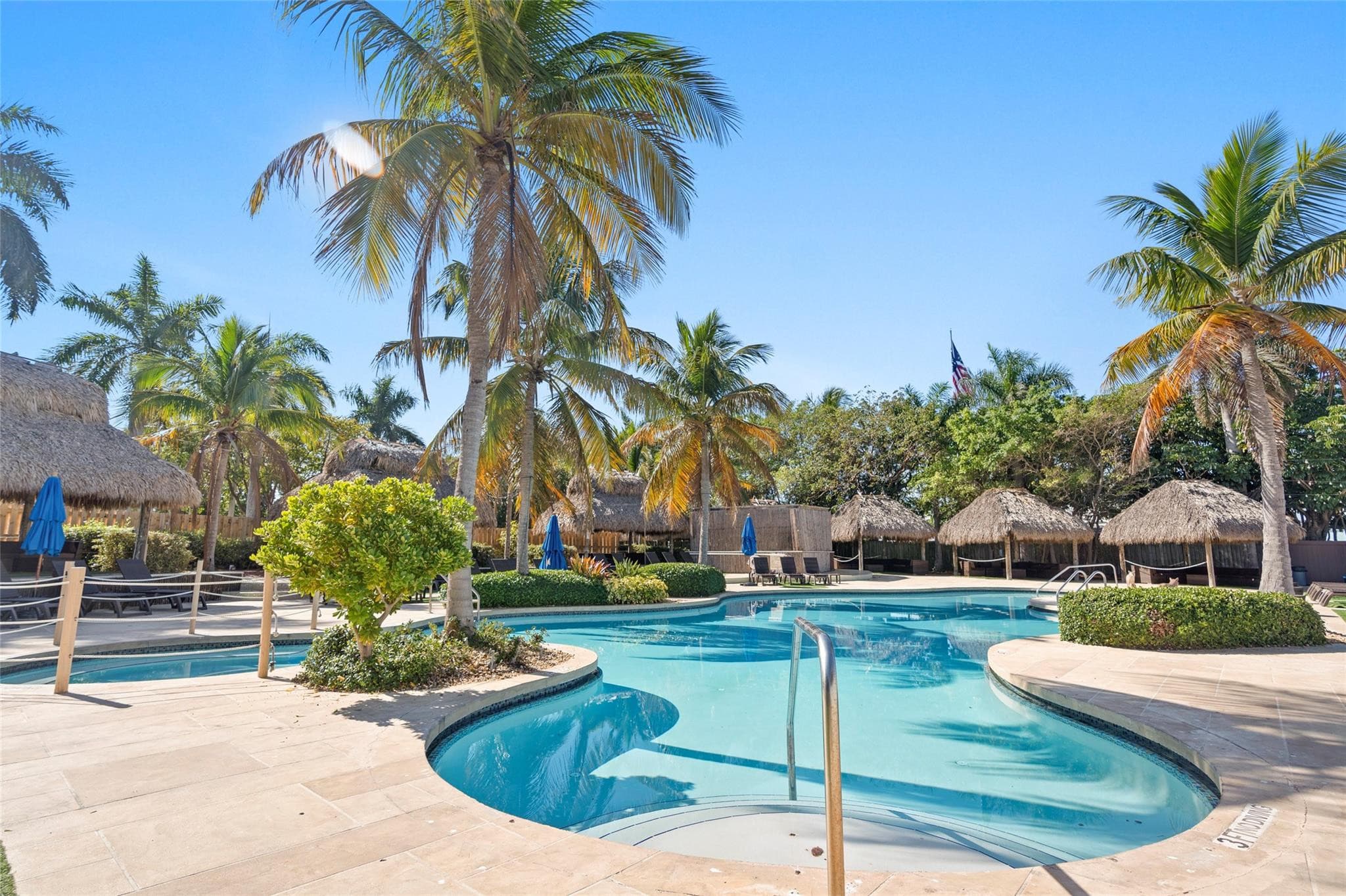 Curved resort pool ringed by travertine decking, tall coconut palms, thatched-roof cabanas, and blue umbrellas under a clear sky.