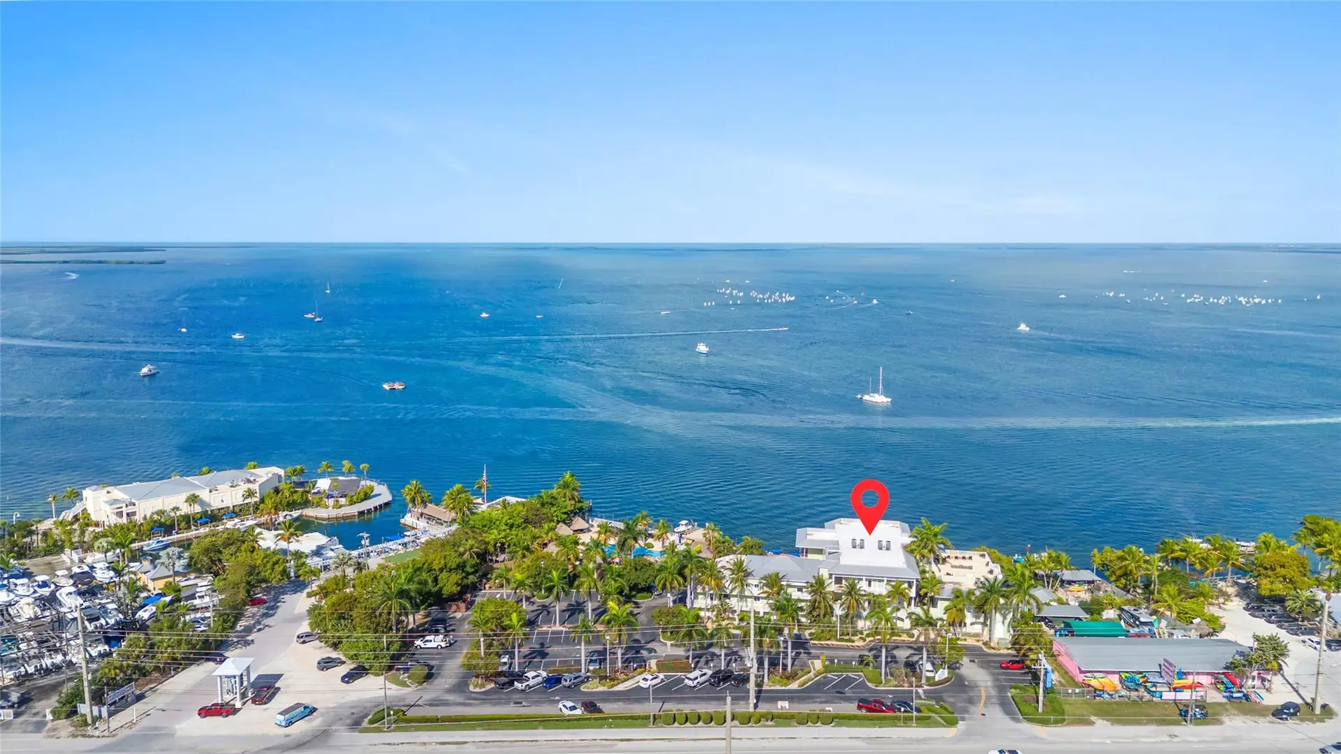 High aerial over the vast blue bay with sailboats, a red location pin marking the white-roofed bayfront residence building amid palms, docks, and the pool below.